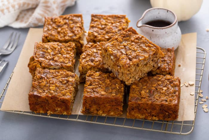 Slices of pumpkin baked oatmeal next to a small pitcher of maple syrup on a piece of parchment paper.