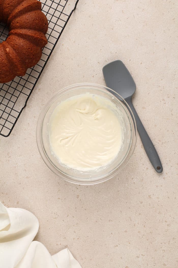 Cream cheese frosting in a glass mixing bowl, set next to a cooling vanilla bundt cake.