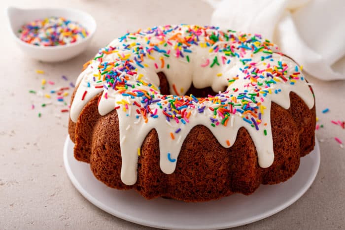 Side view of a vanilla bundt cake topped with cream cheese frosting and sprinkles, set on a white cake plate.