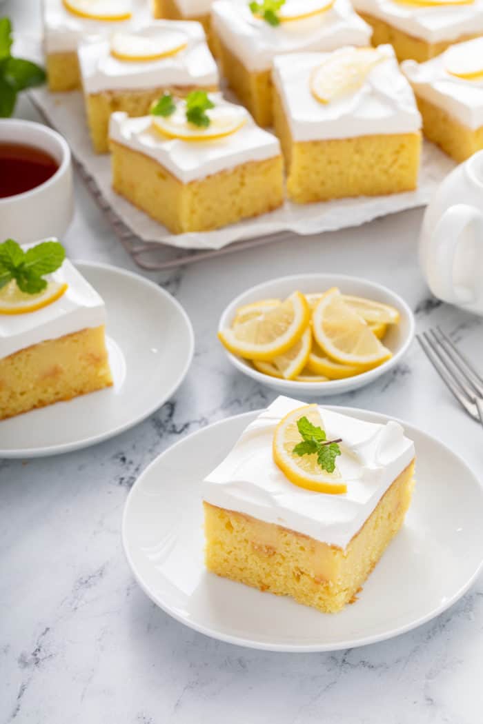 Two white plates holding slices of lemon poke cake, with a platter of the cake in the background.