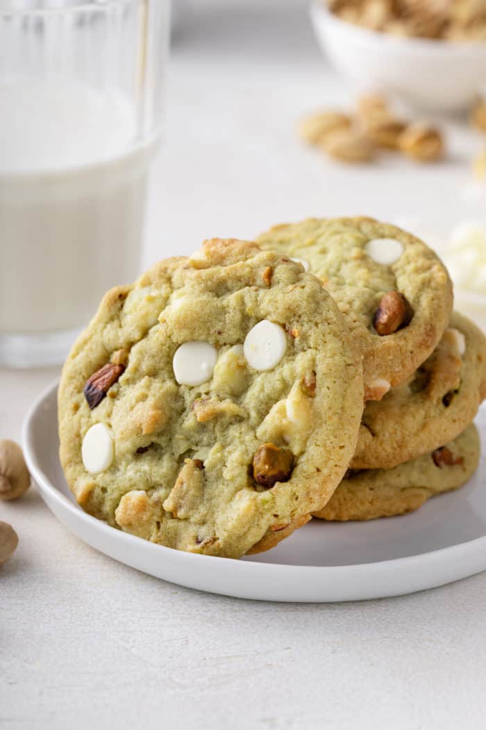 Three white chocolate pistachio pudding cookies stacked on a white plate. A fourth cookie is leaning against the stack.