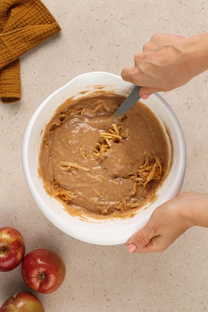 Grated apple being folded into apple butter cake batter in a white mixing bowl.