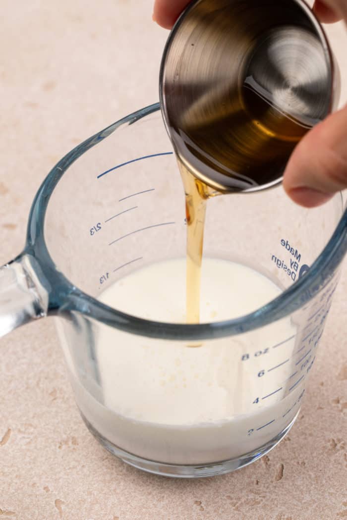 Vanilla syrup being added to cream and milk in a glass measuring cup.