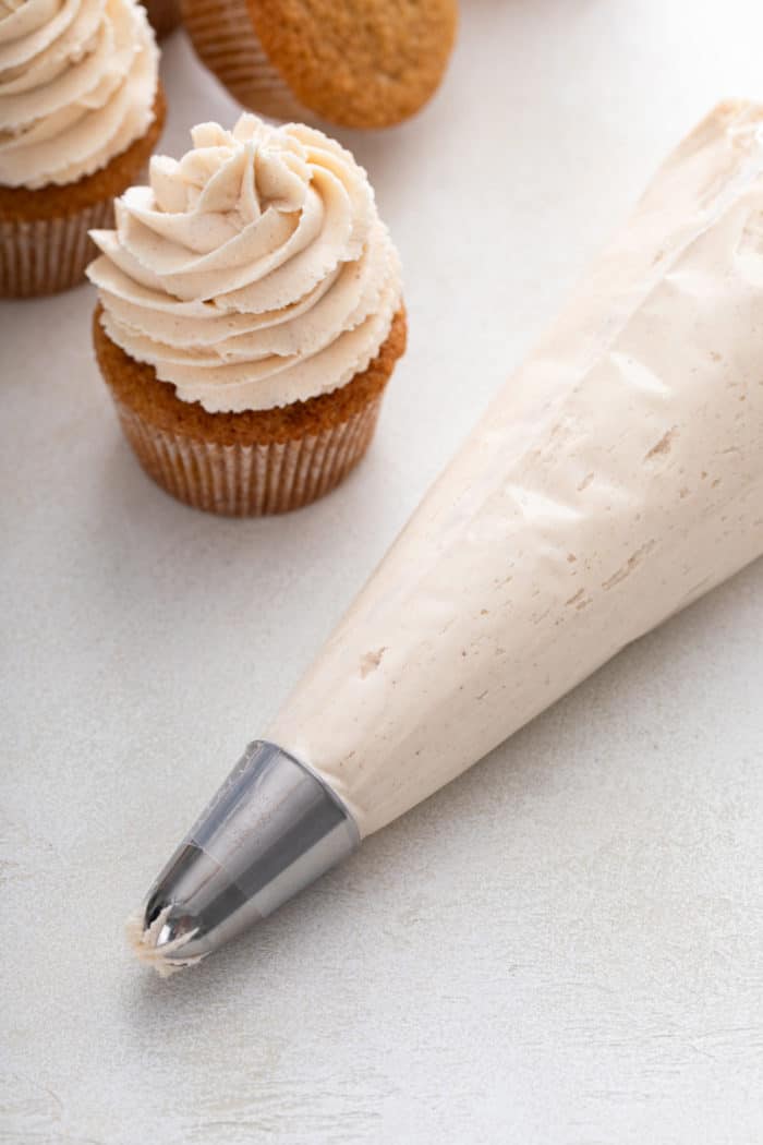 A piping bag filled with brown butter frosting next to a frosted spiced cupcake.