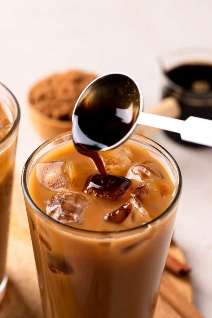 Measuring spoon adding brown sugar syrup to a glass of iced coffee.