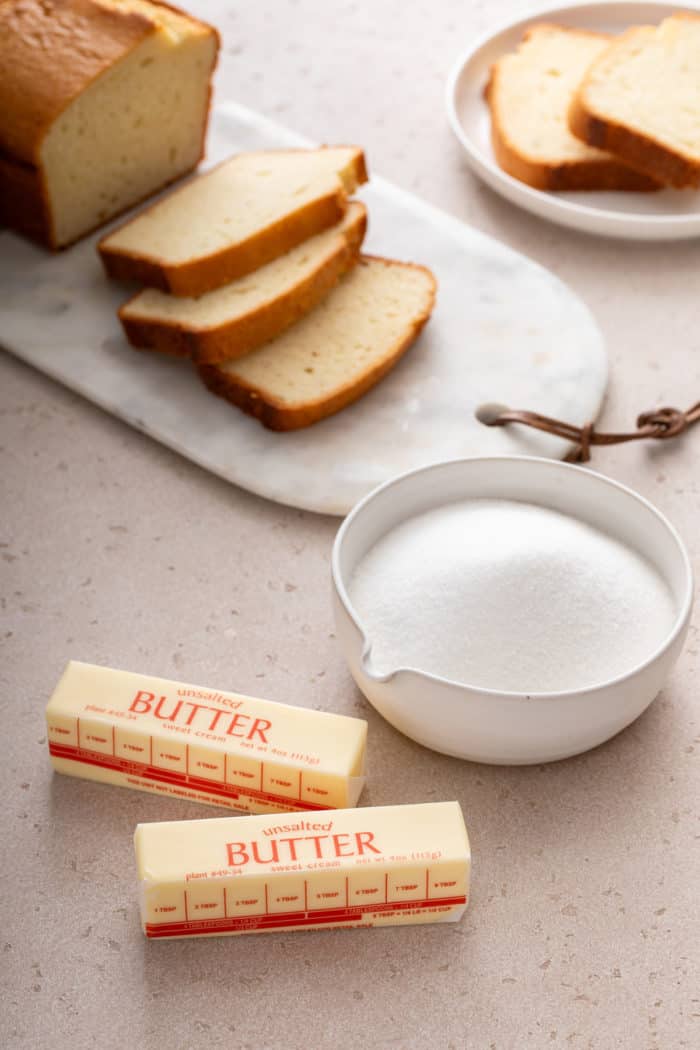 White bowl of sugar and two sticks of butter on a countertop in front of a sliced pound cake.