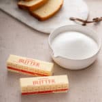 Bowl of sugar and two sticks of butter on a beige countertop.