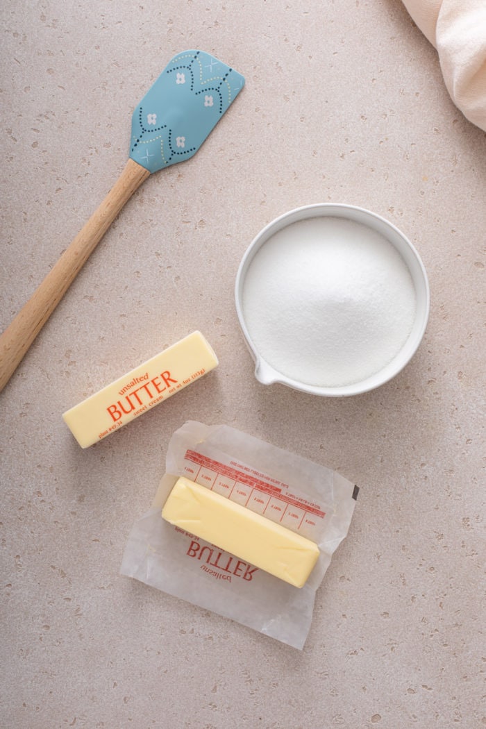 Bowl of sugar, two sticks of butter, and a silicone spatula arranged on a beige countertop. One of the sticks of butter is unwrapped.