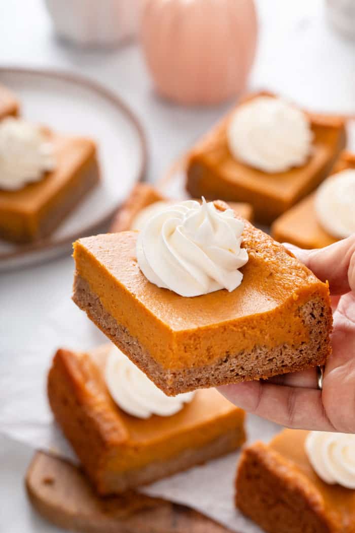 Hand holding up a slice of pumpkin gooey butter cake topped with a dollop of whipped cream.