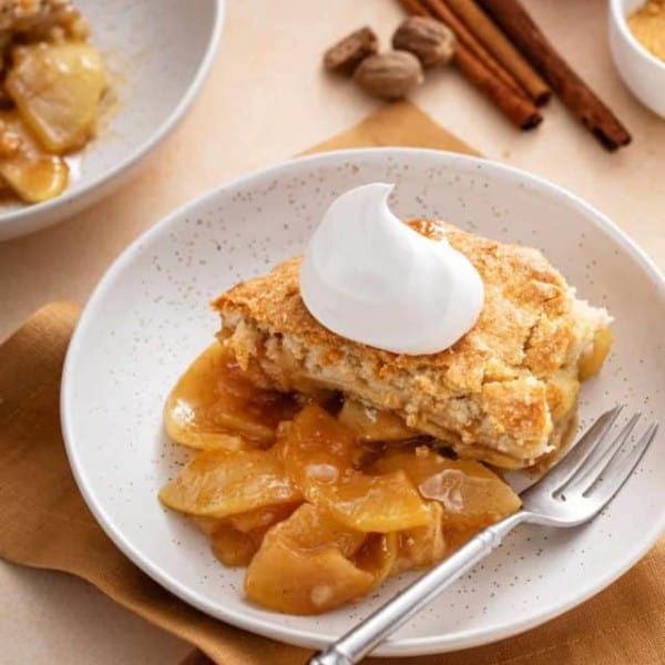 White bowl with a helping of apple cobbler topped with a dollop of whipped cream.