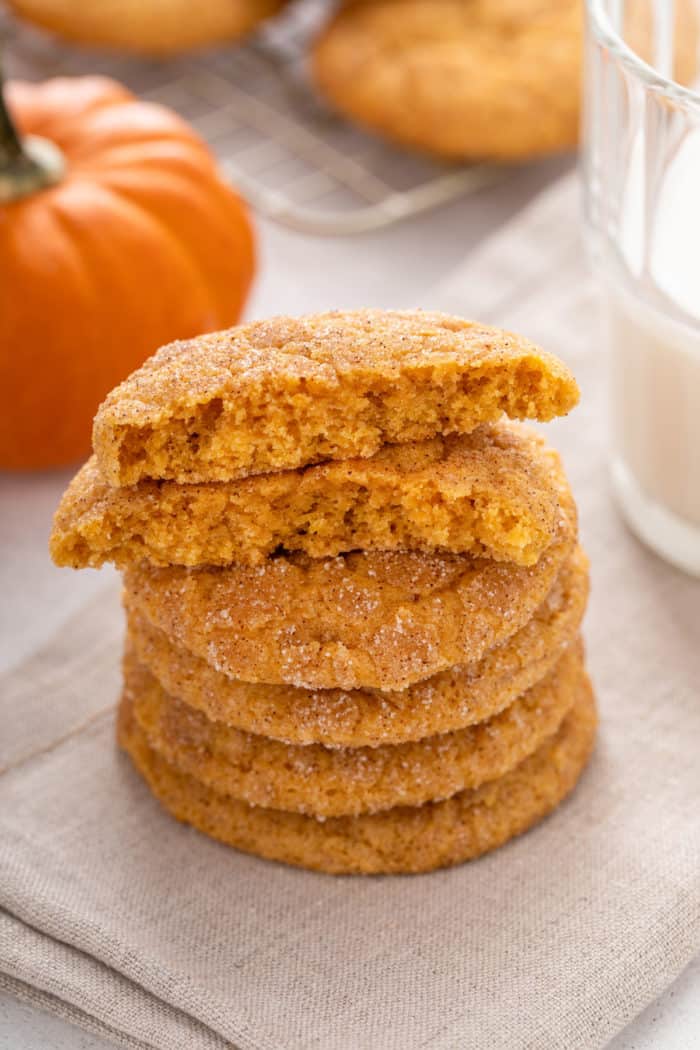 Four pumpkin snickerdoodles stacked on a linen napkin. The top cookie is broken in half to show the inner texture.