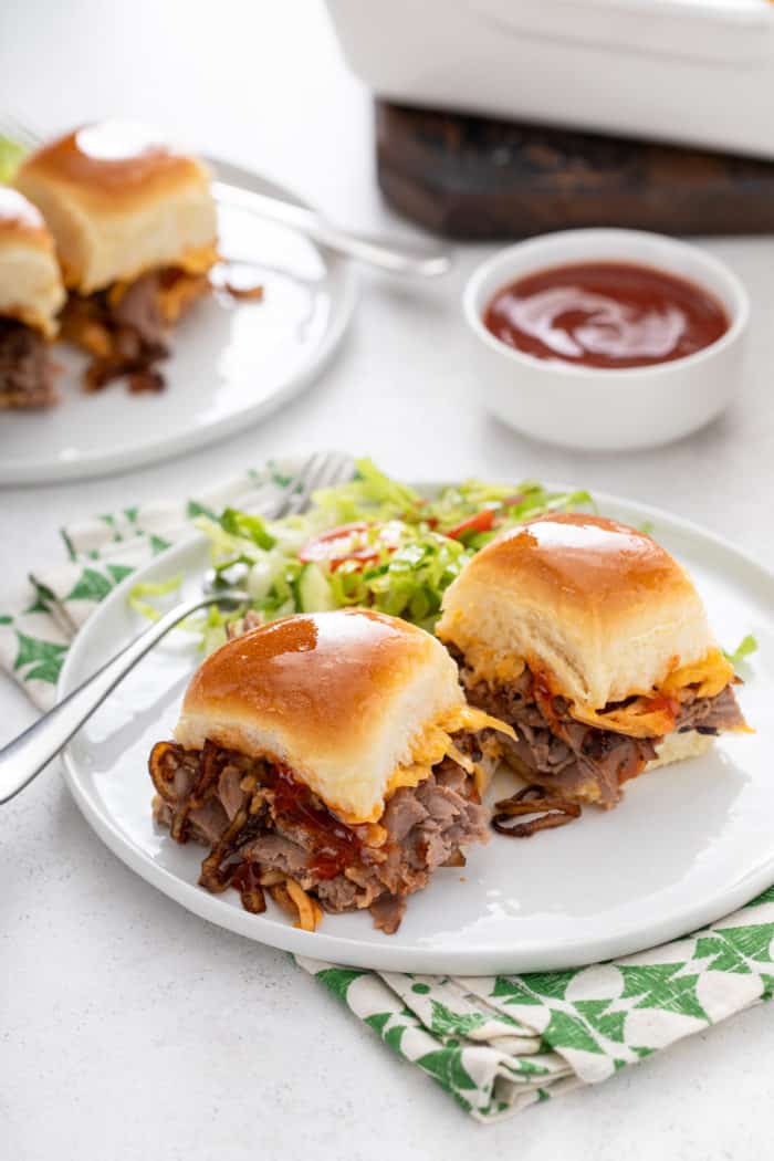 Two cheesy roast beef sliders next to a green salad on a white plate.