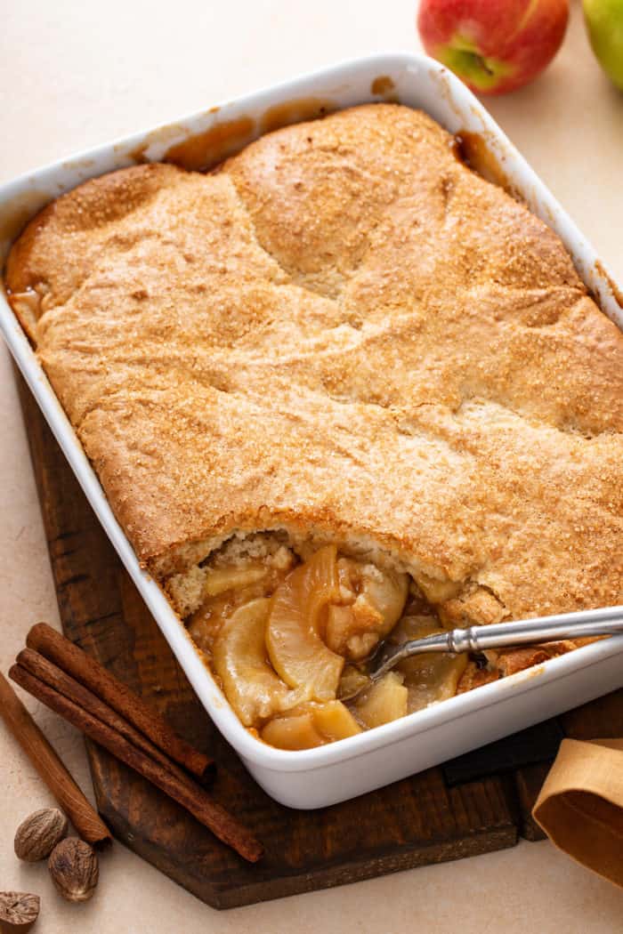 Spoon about to dish out a serving of apple cobbler from a white baking dish.