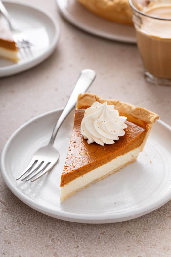 Slice of pumpkin cream cheese pie topped with whipped cream next to a fork on a white plate.