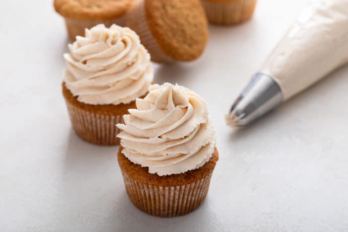 Two cupcakes covered in brown buttercream frosting next to a piping bag filled with frosting.