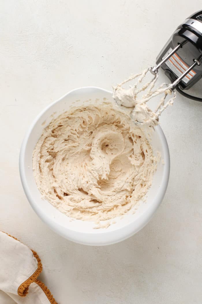 Brown butter frosting beaten in a white mixing bowl next to a hand mixer.