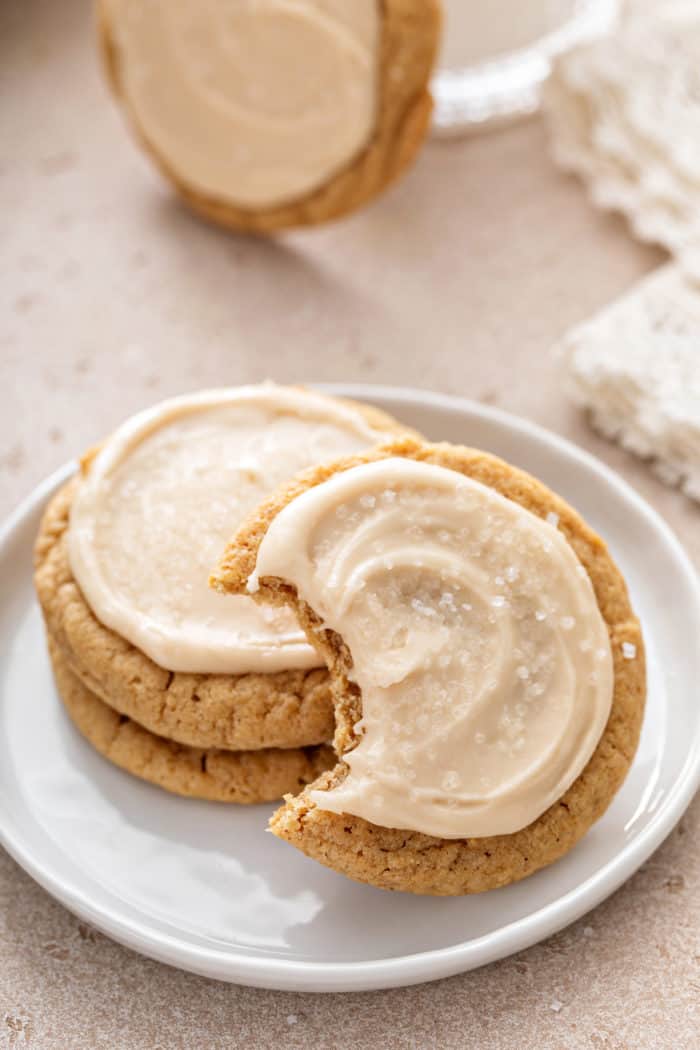 Two stacked maple cookies on a white plate with a third cookie leaning against the stack. A bite has been taken from the third cookie.