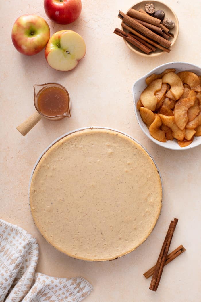 Overhead view of chilled caramel apple cheesecake next to a bowl of cooked apples and a jar of caramel sauce.