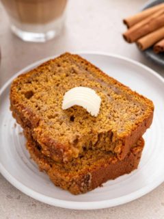 Two slices of pumpkin banana bread on a white plate. The top slice has a pat of butter and a bite taken from the corner.