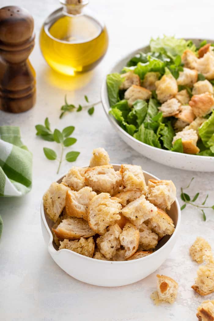 Small white bowl of croutons on a countertop with a bowl of salad in the background.