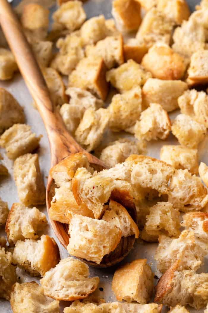 Wooden spoon holding a spoonful of homemade croutons on a baking sheet.