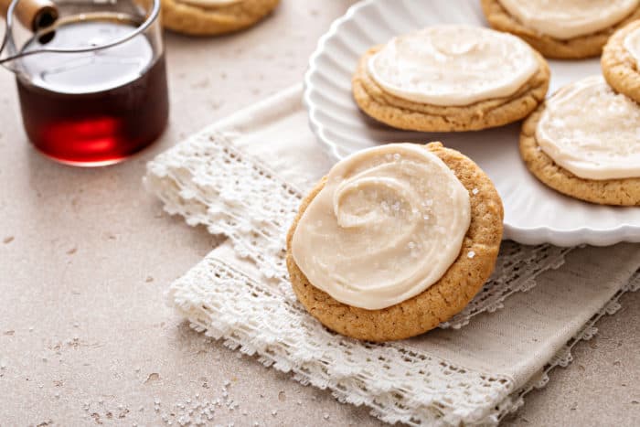 Frosted maple cookie leaning against a white plate of more cookies.