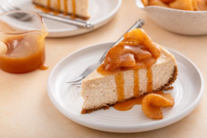 Slice of caramel apple cheesecake on a white plate. A jar of caramel is visible in the background.