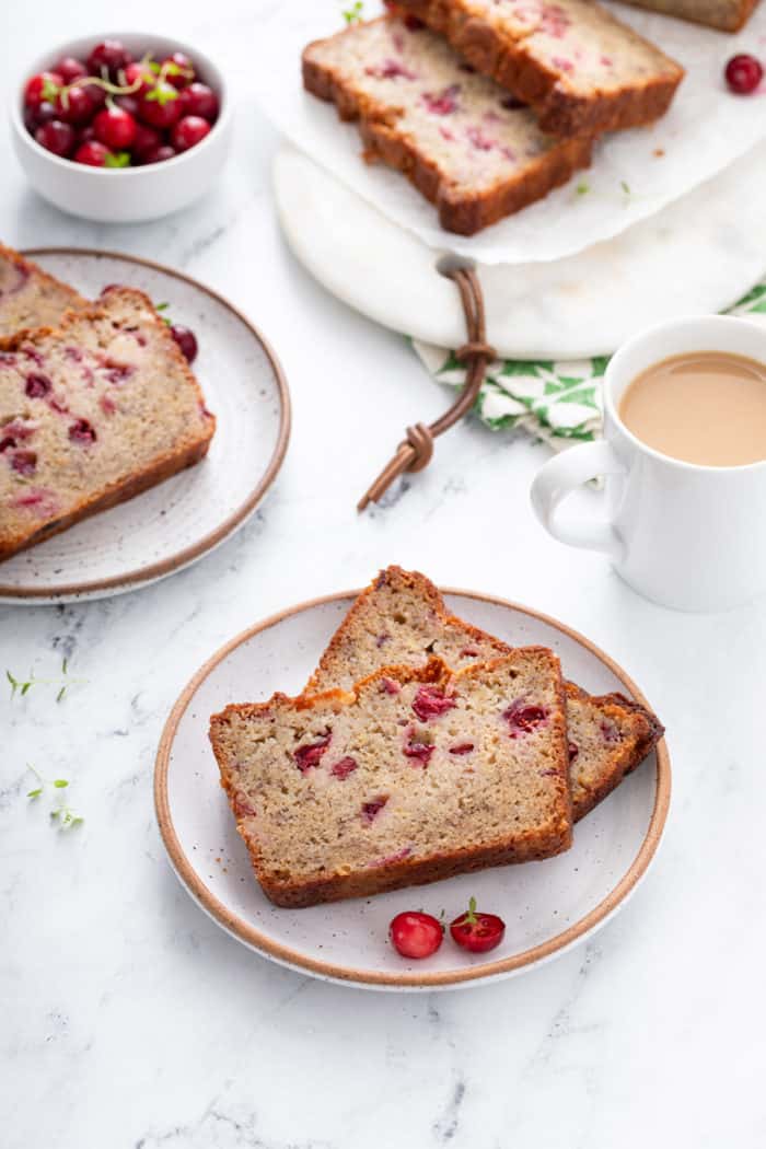 Two plates, each holding two slices of cranberry banana bread, with a loaf of the bread in the background.