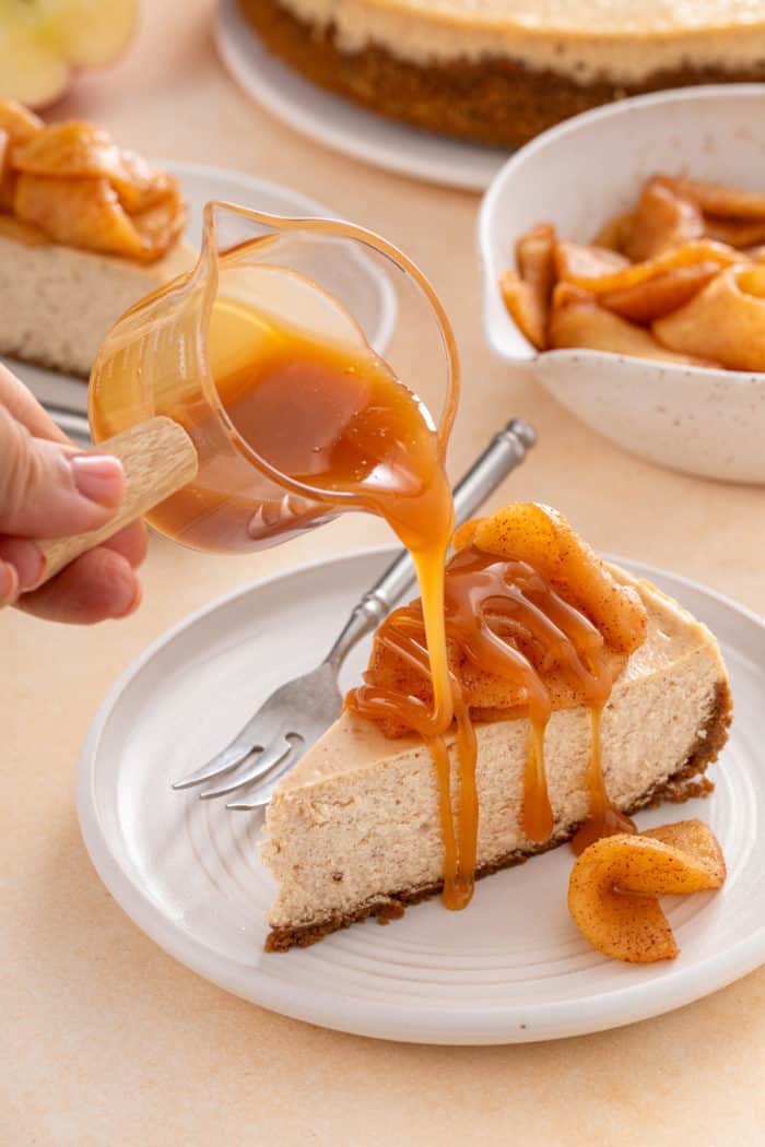 Caramel sauce is being poured over a slice of caramel apple cheesecake on a white plate.