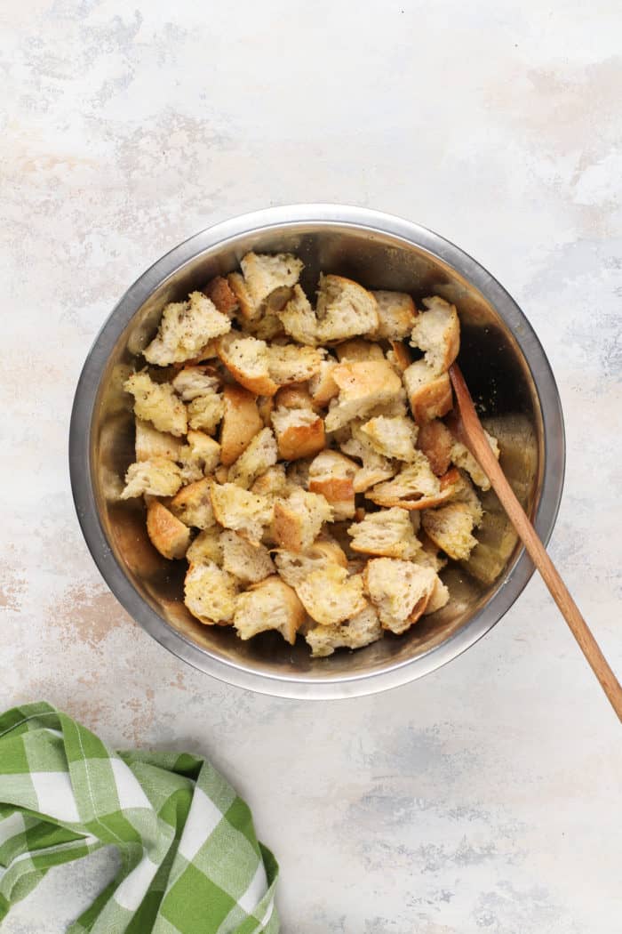 Wooden spoon tossing cubes of bread with seasoning in a metal bowl.