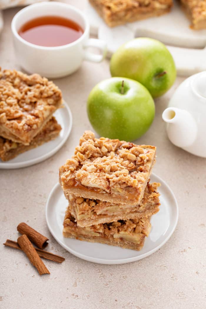 Three apple crisp bars stacked on a white plate with green apples and more bars in the background.