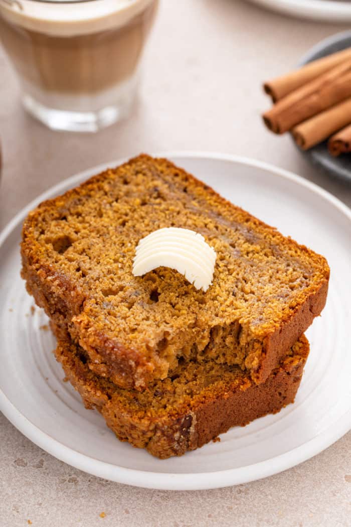 Two slices of pumpkin banana bread on a white plate. The top slice has a pat of butter and a bite taken from the corner.