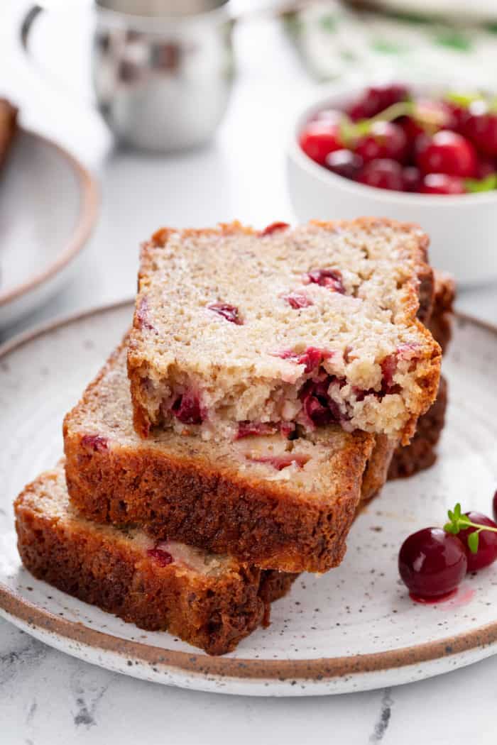 Stacked slices of cranberry banana bread on a speckled plate. The top slice is broken in half to show the crumb.