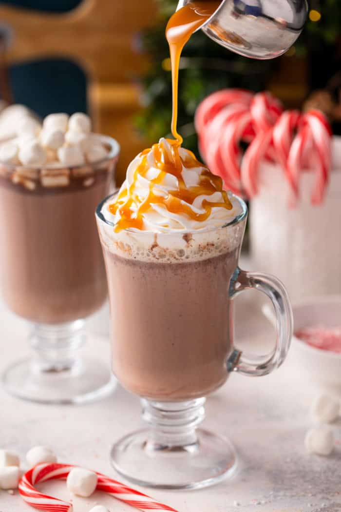 Salted caramel sauce being drizzled over whipped-cream-topped hot chocolate in a glass mug.