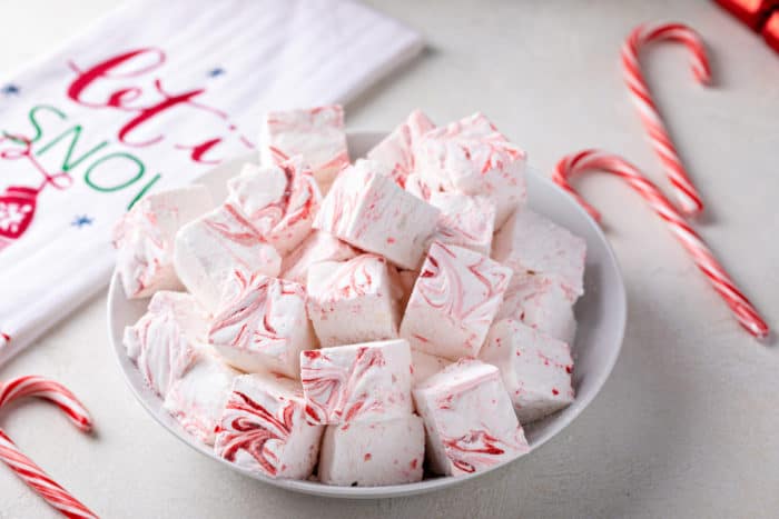 White bowl filled with peppermint marshmallows on a countertop next to candy canes and a festive tea towel.
