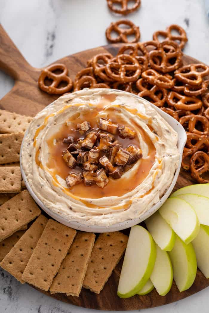 Bowl of snickers dip on a wooden board surrounded by graham crackers, sliced apples, and pretzel twists.