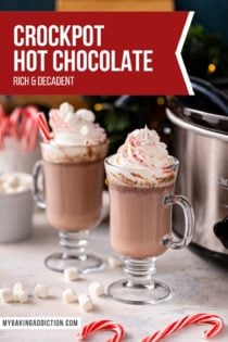 Two glass mugs of crockpot hot chocolate topped with marshmallows and whipped cream. The slow cooker is visible in the background. Text overlay includes recipe name.