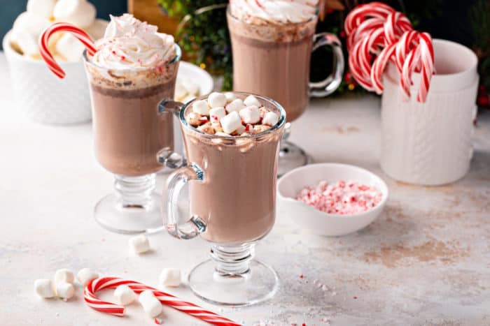 Three glass mugs of crockpot hot chocolate on a countertop next to bowls of marshmallows and candy canes.