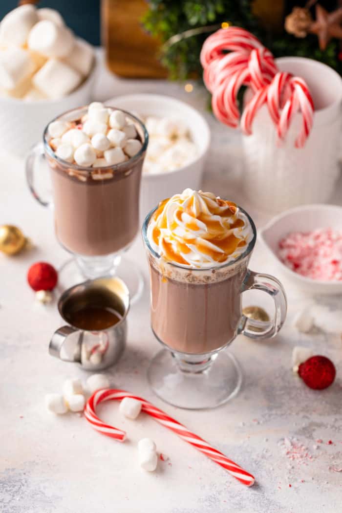 Two glass mugs filled with crockpot hot chocolate and surrounded by various toppings, including peppermints, marshmallows, and caramel sauce.