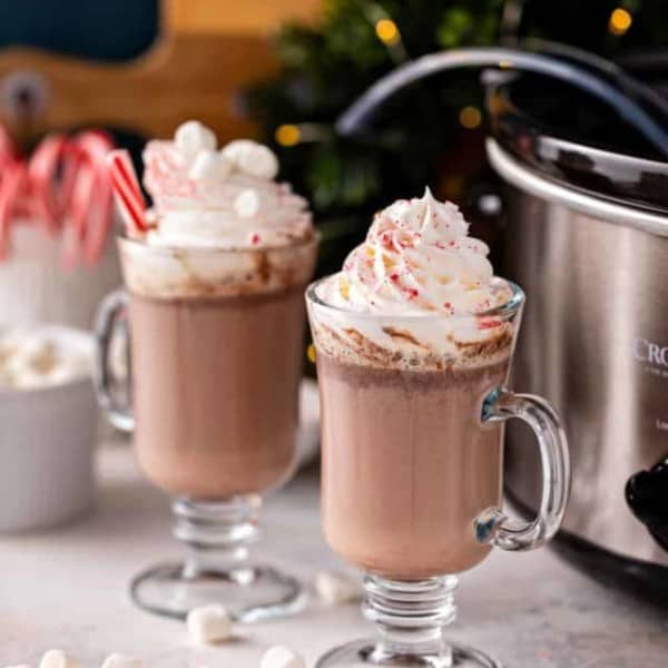 Two glass mugs of crockpot hot chocolate topped with marshmallows and whipped cream. The slow cooker is visible in the background.