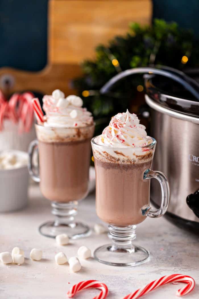 Two glass mugs of crockpot hot chocolate topped with marshmallows and whipped cream. The slow cooker is visible in the background.