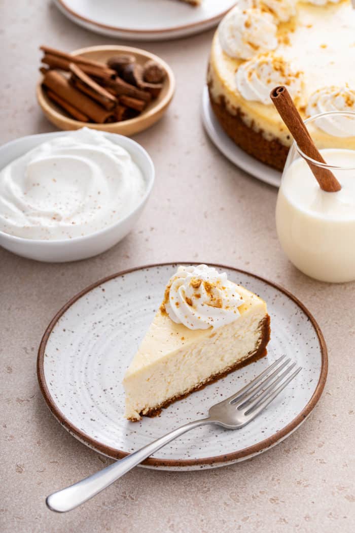 Slice of eggnog cheesecake on a stoneware plate with a glass of eggnog and a bowl of whipped cream in the background.