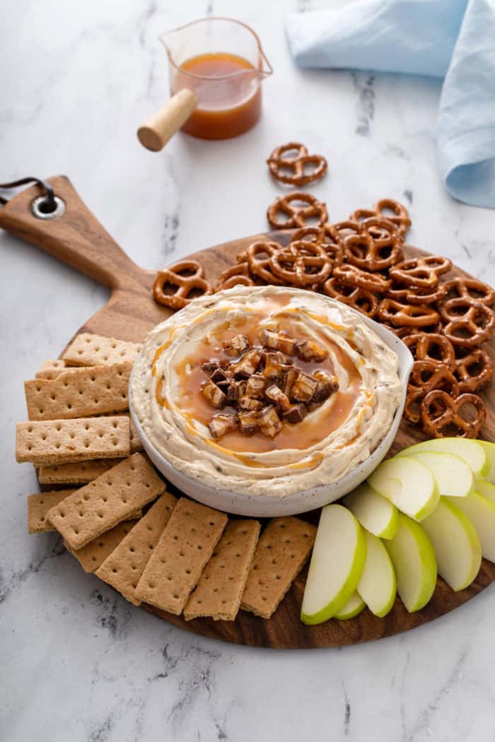 Wooden board arranged with a bowl of snickers dip, graham crackers, apples, and pretzels.