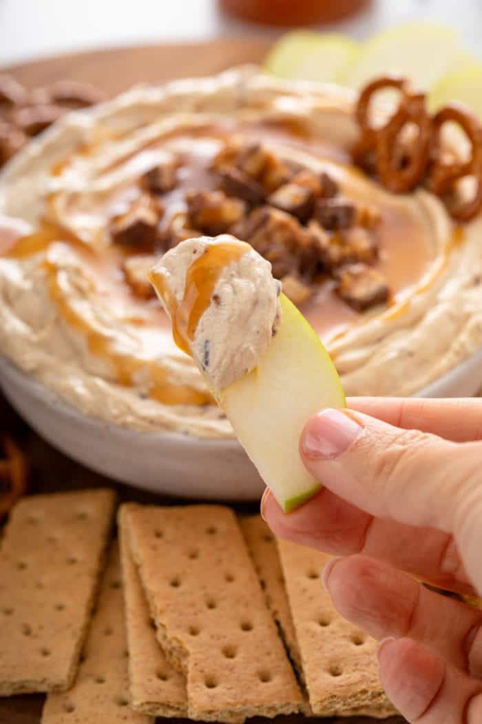 Hand holding up a slice of apple with snickers dip on it. A bowl of the dip is visible in the background.