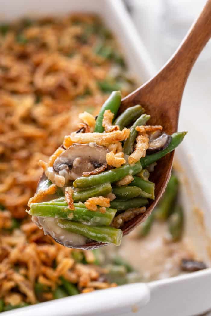 Wooden spoon holding up fresh green bean casserole to the camera, with the filled baking dish in the background.