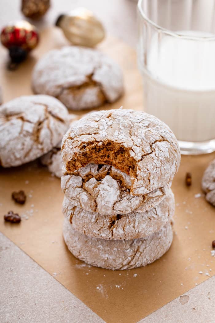 Four gingerbread crinkle cookie stacked in front of a glass of milk. The top cookie in the stack has a bite taken from it.