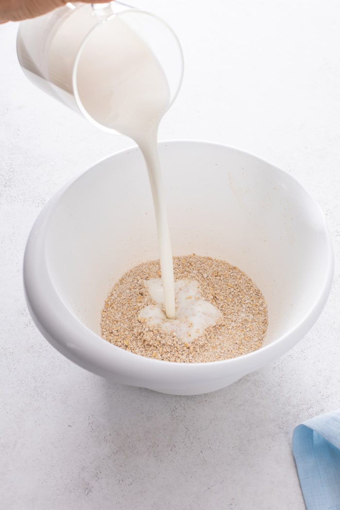 Buttermilk being poured over oats in a white mixing bowl.