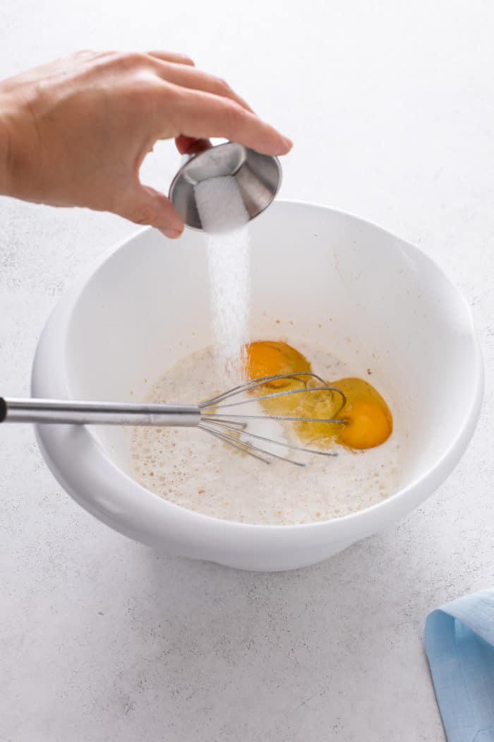 Eggs and sugar being added to oats and buttermilk in a white bowl.