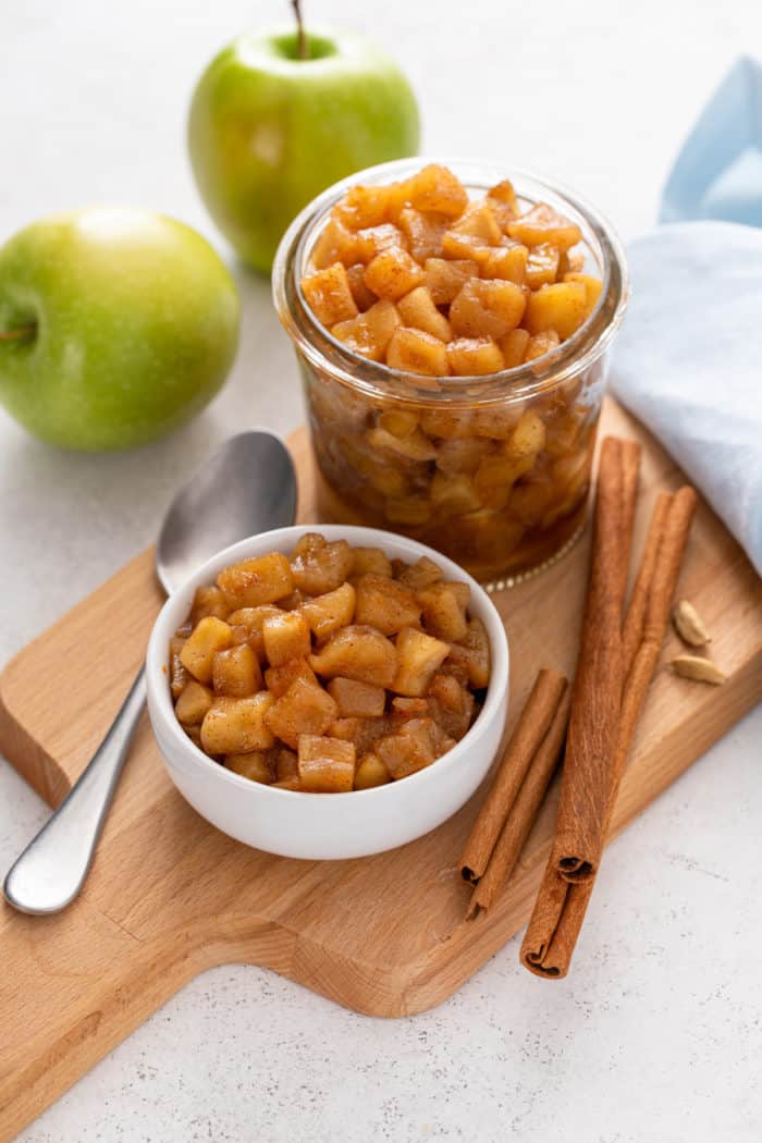 Glass jar and white bowl, both filled with apple compote and set on a wooden board next to cinnamon sticks.