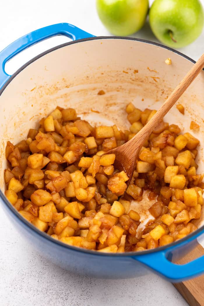 Cooked apple compote in a blue dutch oven being stirred with a wooden spoon.